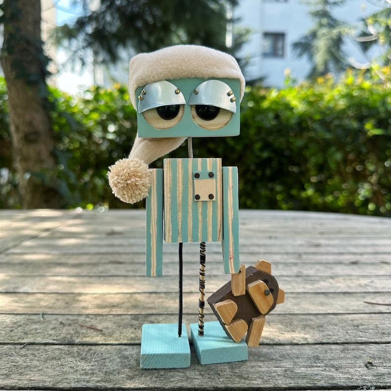 May include: A blue and white striped wooden robot wearing a white knit hat with a pom-pom. The robot has large, round, black eyes and is holding a small, brown wooden animal.