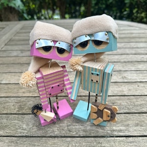 May include: Two wooden robot figurines wearing sleep caps. The robot on the left is pink and white striped with a small doll in its hand. The robot on the right is blue and white striped and holding a small wooden animal.