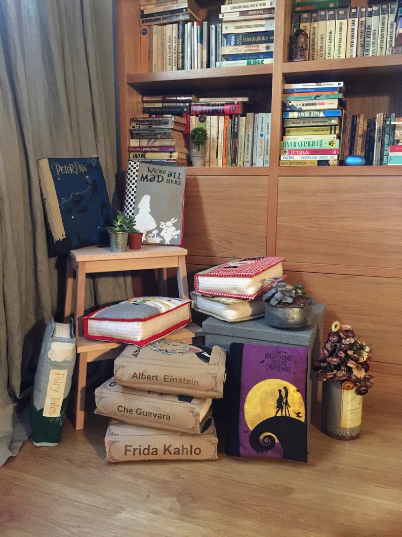 May include: A collection of book-shaped decorative pillows with various titles, including "Peter Pan", "We're All Mad Here", "Fairy Tales", "Albert Einstein", "Che Guevara", and "Frida Kahlo". The pillows are arranged on a wooden floor with a bookshelf in the background.