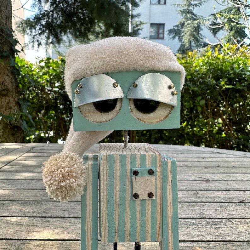 May include: A wooden robot wearing a white fuzzy hat with a pom-pom. The robot has a light blue body with white stripes, silver eye covers, and brown eyes.