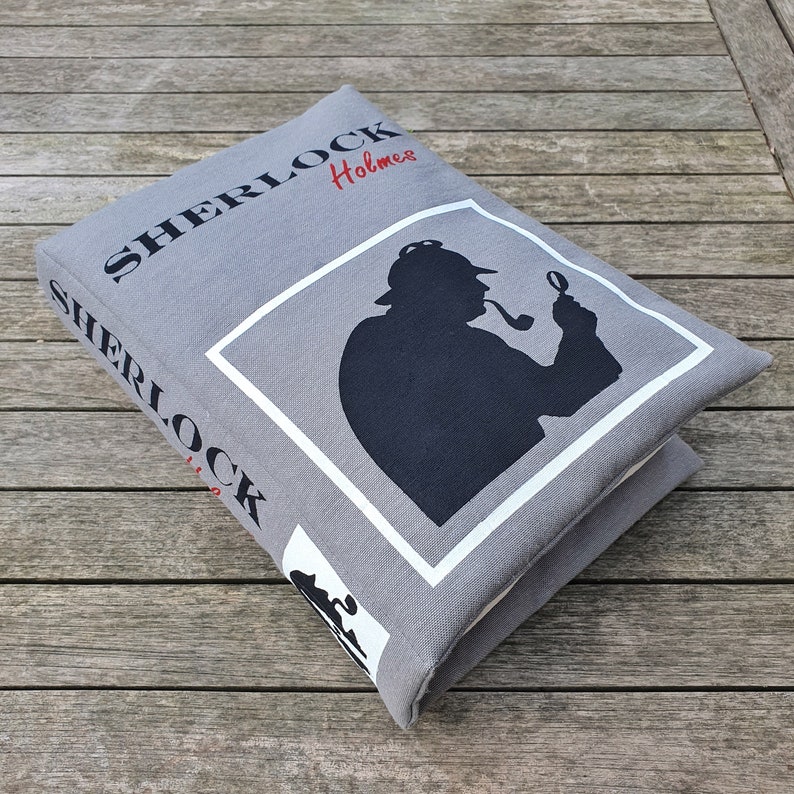 May include: A gray canvas book with the text "SHERLOCK Holmes" printed in black. The book features a silhouette of a person wearing a hat and holding a magnifying glass and pipe.