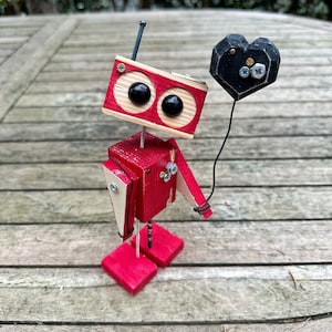 May include: A red and white wooden robot holding a black heart-shaped object. The robot has two large black eyes and a small antenna on top of its head.