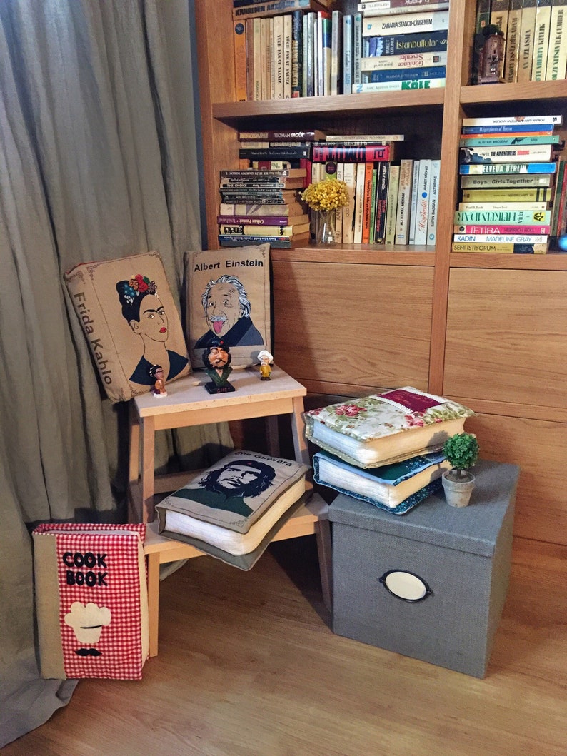 May include: A bookshelf with various books, including a cook book, and decorative pillows with images of Frida Kahlo, Albert Einstein, and Che Guevara. The pillows are sitting on a wooden stool and a gray storage box.