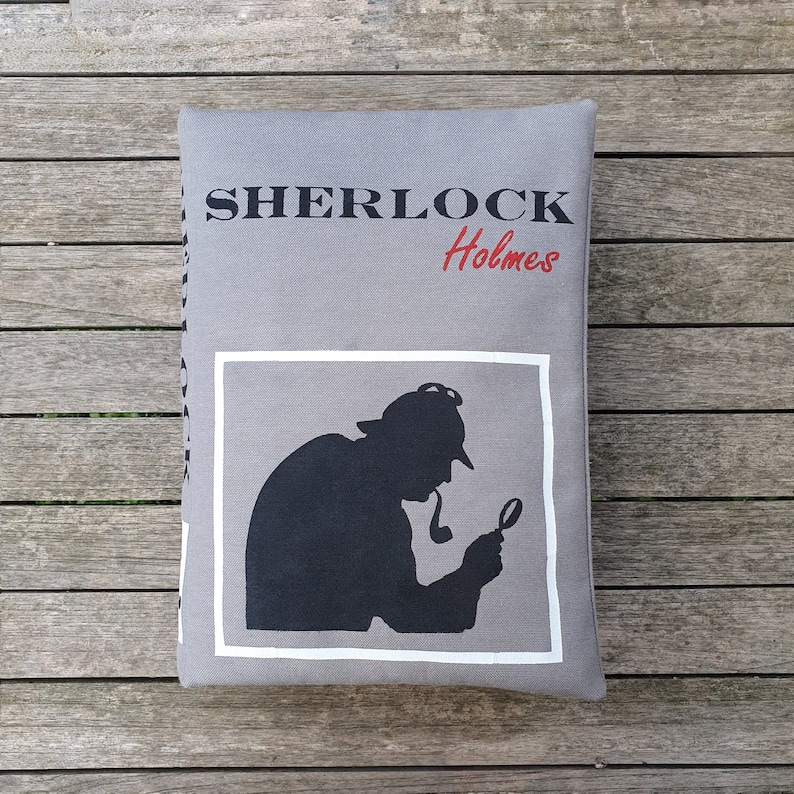 May include: A gray pillow with the text "SHERLOCK Holmes" printed in black and red. The pillow features a black silhouette of a man with a magnifying glass.