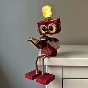 Puede incluir: Una figurita de robot de madera con una cabeza de bombilla, leyendo un libro y sentada en un estante blanco. El robot est&aacute; pintado de rojo y tiene grandes ojos negros redondos.