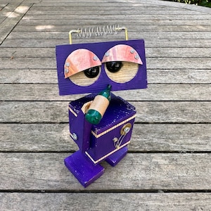 May include: A purple wooden robot with a spring on its head, holding a green bottle in its hand. The robot has large, round eyes and a whimsical design.