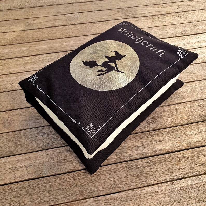 May include: Black and white pillow with a witch flying on a broomstick in front of a full moon. The pillow has a white border and the text "Witchcraft" in white lettering.