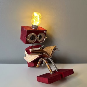 Puede incluir: Un robot de madera rojo con una cabeza de bombilla, que lleva gafas y lee un libro. El robot tiene piernas de alambre de cobre y est&aacute; sentado sobre una superficie blanca.