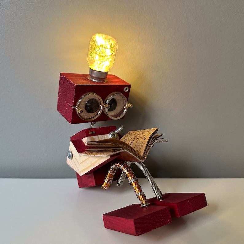 Puede incluir: Un robot de madera rojo con una cabeza de bombilla, que lleva gafas y lee un libro. El robot tiene piernas de alambre de cobre y est&aacute; sentado sobre una superficie blanca.