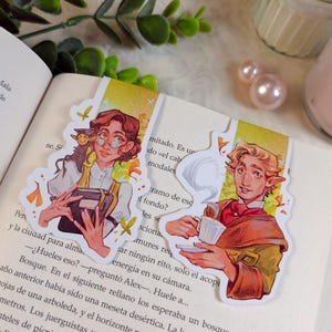 Puede incluir: Dos marcapáginas ilustrados con personajes con libros y una taza de té. Los marcapáginas tienen un fondo amarillo y blanco con un diseño floral. Los personajes llevan gafas y una chaqueta roja.