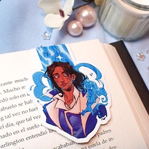 May include: A blue and white bookmark featuring a cartoon illustration of a man with long dark hair and a blue and white jacket. The man is surrounded by blue and white swirls. The bookmark is placed on a book with text in Spanish.