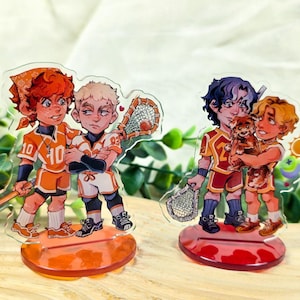 Puede incluir: Dos figuras de acrílico con personajes de dibujos animados con uniformes deportivos. Una figura muestra dos figuras con palos y redes de lacrosse, la otra muestra dos figuras sosteniendo un perro pequeño. Las figuras están sobre bases naranjas y rojas.