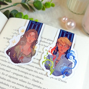 May include: Two illustrated bookmarks featuring fantasy characters. One bookmark depicts a woman with long brown hair, and the other shows a man with blonde hair. Both bookmarks have a decorative blue and gold border and are placed on the pages of a book.