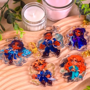 Puede incluir: Cinco llaveros de personajes coloridos con fondos transparentes. Cada llavero presenta un personaje diferente con un diseño y una pose únicos. Los personajes llevan todos atuendos azules y blancos y tienen diferentes colores de cabello. Los llaveros cuelgan de anillos de metal dorado.