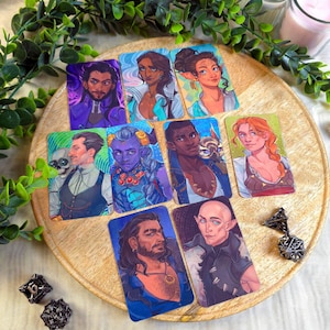 Puede incluir: Una colección de tarjetas de arte de personajes con retratos de fantasía. Las tarjetas muestran diversos personajes con rasgos faciales, ropa y accesorios detallados. Las tarjetas están dispuestas sobre una bandeja de madera con dados decorativos.