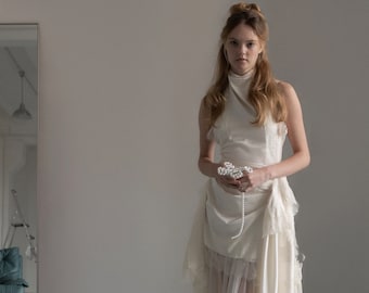 May include: Ivory-colored bridal gown with a high neckline and layered skirt. The dress features a long train and is paired with delicate sandals. A pearl necklace and a floral accessory are held in the hands.