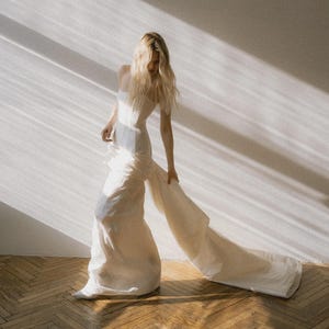 Peut inclure: Une robe longue crème avec un corsage ajusté et une jupe fluide. La robe a une longue traîne et est portée par une personne aux longs cheveux blonds. Le fond est un mur blanc avec la lumière du soleil et un sol en bois.