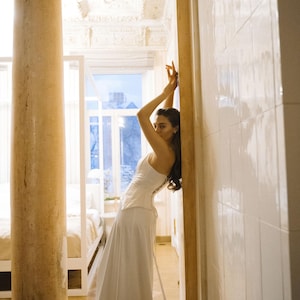May include: A white bridal gown with a fitted bodice and a flowing skirt. The bodice features a corset-style back. The gown is made of a lightweight, flowing fabric. The bride is posing in a luxurious, ornate room with tall columns and high ceilings. The overall theme is bridal fashion and wedding attire.