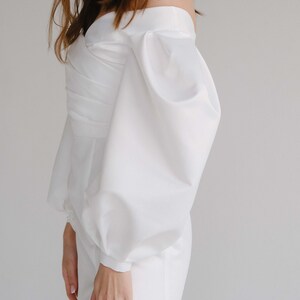 Puede incluir: Un mono blanco con hombros descubiertos y mangas voluminosas. El mono presenta un corpi&ntilde;o ajustado con un dise&ntilde;o cruzado y un dobladillo corto. Las mangas est&aacute;n recogidas en la mu&ntilde;eca, creando un efecto globo. La tela parece ser lisa, posiblemente sat&eacute;n.