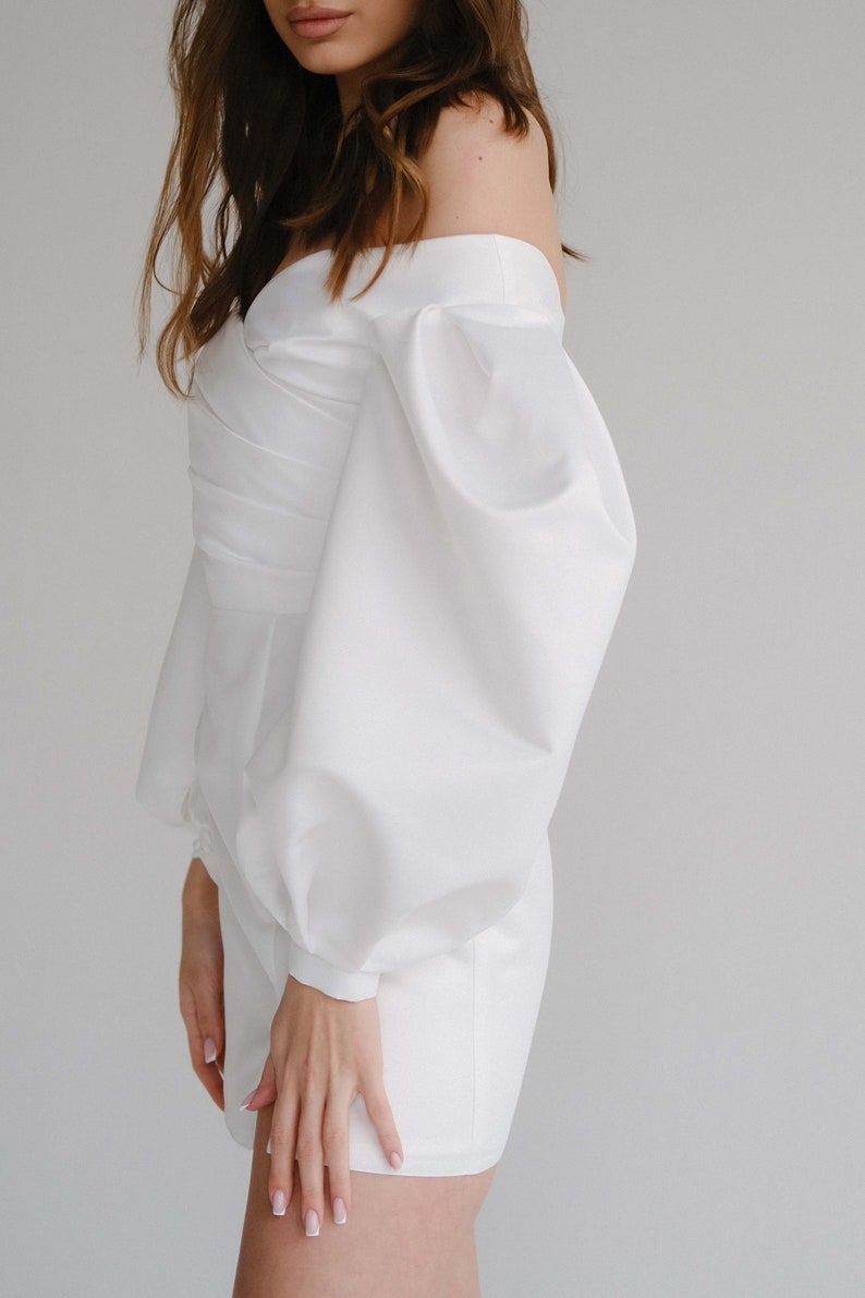 Puede incluir: Un mono blanco con hombros descubiertos y mangas voluminosas. El mono presenta un corpi&ntilde;o ajustado con un dise&ntilde;o cruzado y un dobladillo corto. Las mangas est&aacute;n recogidas en la mu&ntilde;eca, creando un efecto globo. La tela parece ser lisa, posiblemente sat&eacute;n.