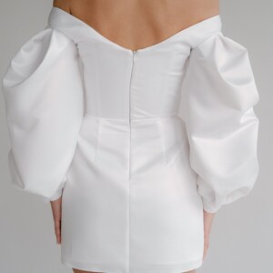 Puede incluir: Un minivestido blanco con hombros descubiertos y mangas abullonadas voluminosas. El vestido presenta un corpi&ntilde;o ajustado y una falda corta, con cierre de cremallera en la espalda. La tela tiene un ligero brillo, lo que sugiere sat&eacute;n o un material similar.