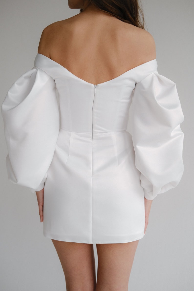 Puede incluir: Un minivestido blanco con hombros descubiertos y mangas abullonadas voluminosas. El vestido presenta un corpi&ntilde;o ajustado y una falda corta, con cierre de cremallera en la espalda. La tela tiene un ligero brillo, lo que sugiere sat&eacute;n o un material similar.