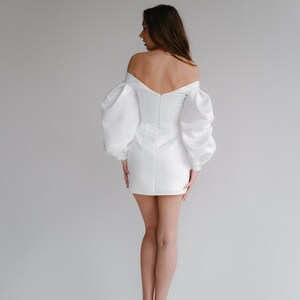 Puede incluir: Un minivestido blanco con escote de hombros descubiertos y mangas abullonadas voluminosas. El vestido presenta un corpi&ntilde;o ajustado y una falda corta. La modelo lleva tacones color nude. El vestido es adecuado para una boda u ocasi&oacute;n especial.