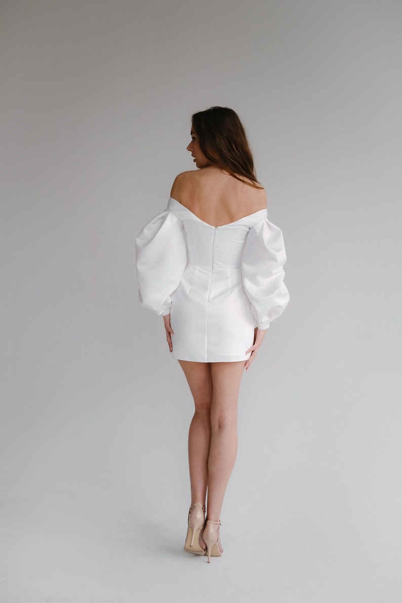 Puede incluir: Un minivestido blanco con escote de hombros descubiertos y mangas abullonadas voluminosas. El vestido presenta un corpi&ntilde;o ajustado y una falda corta. La modelo lleva tacones color nude. El vestido es adecuado para una boda u ocasi&oacute;n especial.