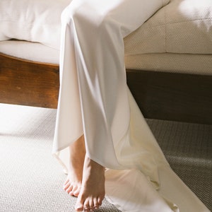 May include: A white satin dress with a long train, draped over a wooden bed frame. The dress is gathered at the waist and falls to the floor in soft folds.