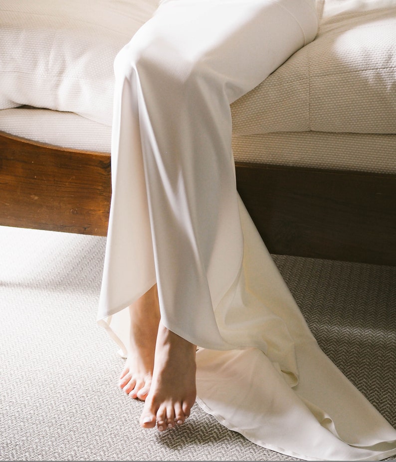 May include: A white satin dress with a long train, draped over a wooden bed frame. The dress is gathered at the waist and falls to the floor in soft folds.