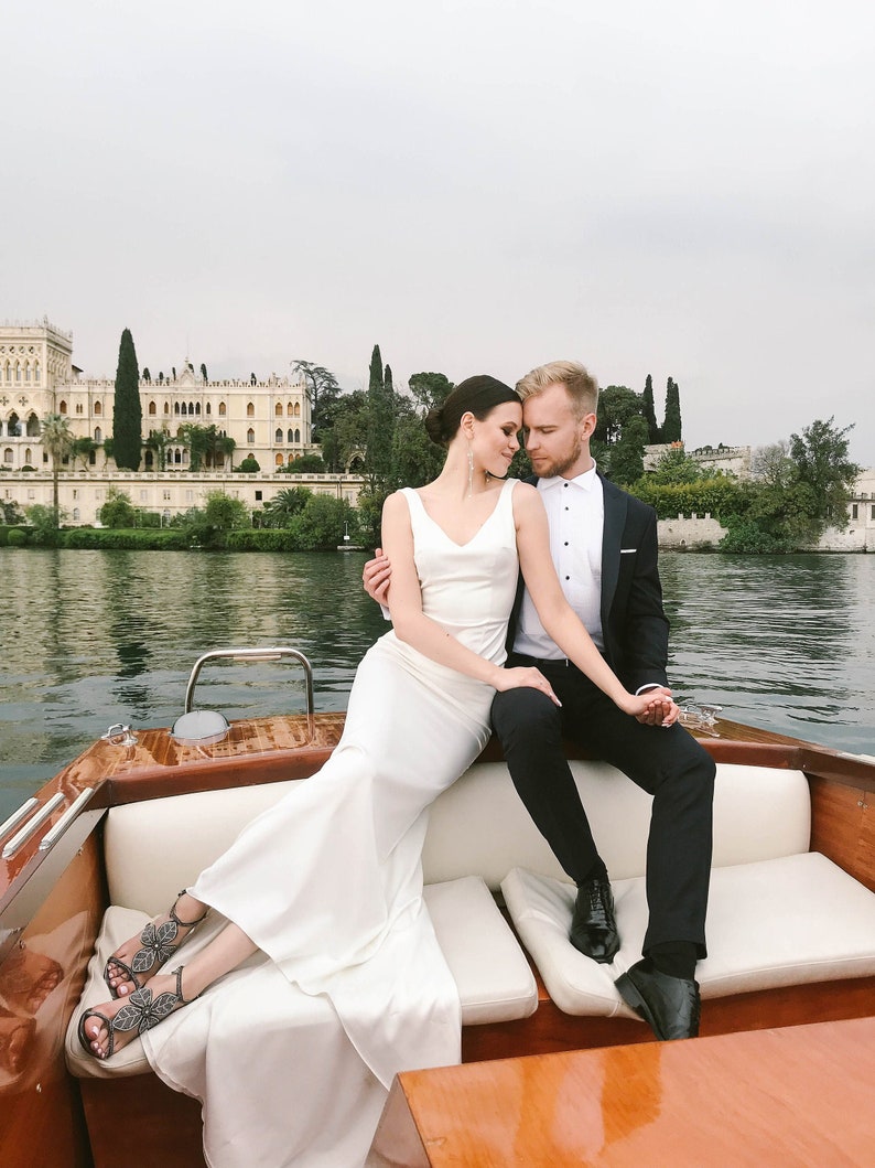 May include: A bride and groom in a white wedding dress and a black suit are sitting on a wooden boat. The bride is wearing a white dress with a low neckline and a long train. The groom is wearing a black suit with a white shirt and a black tie. They are looking at each other and smiling. The boat is on a lake with a building in the background.