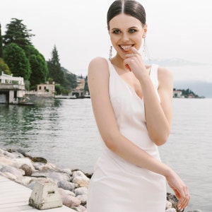 May include: A woman in a white sleeveless dress smiles and touches her chin with her finger. She is standing on a wooden dock with rocks and water in the background.