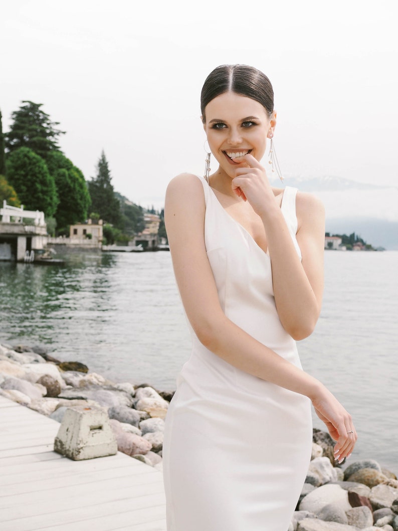 May include: A woman in a white sleeveless dress smiles and touches her chin with her finger. She is standing on a wooden dock with rocks and water in the background.