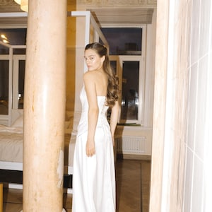 May include: A floor-length white satin wedding gown with a fitted bodice and a flowing skirt. The bodice features a corset-style back. The gown is shown on a model in a room with a marble column and hardwood floors. The overall style is elegant and minimalist.