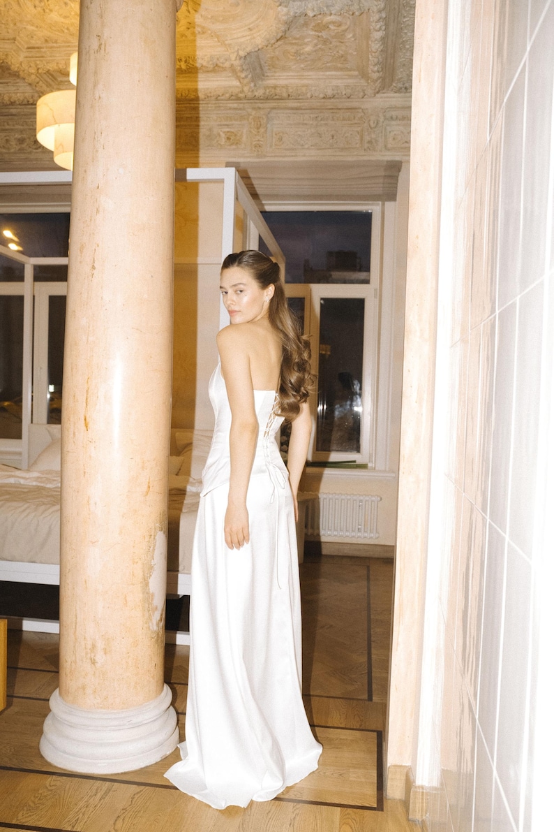 May include: A floor-length white satin wedding gown with a fitted bodice and a flowing skirt. The bodice features a corset-style back. The gown is shown on a model in a room with a marble column and hardwood floors. The overall style is elegant and minimalist.