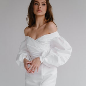 Puede incluir: Un vestido blanco con hombros descubiertos y mangas largas abullonadas. El vestido presenta un corpi&ntilde;o cruzado y una abertura en el muslo. El vestido es usado por una modelo. El vestido est&aacute; hecho de una tela lisa, similar al sat&eacute;n.