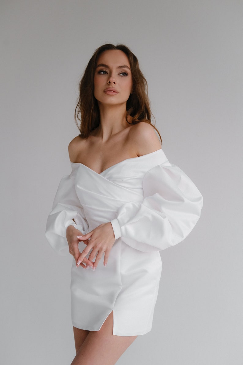 Puede incluir: Un vestido blanco con hombros descubiertos y mangas largas abullonadas. El vestido presenta un corpi&ntilde;o cruzado y una abertura en el muslo. El vestido es usado por una modelo. El vestido est&aacute; hecho de una tela lisa, similar al sat&eacute;n.