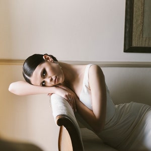 May include: A woman in a white dress is lying on a white couch with her head resting on her arm. She is looking at the camera with a thoughtful expression.