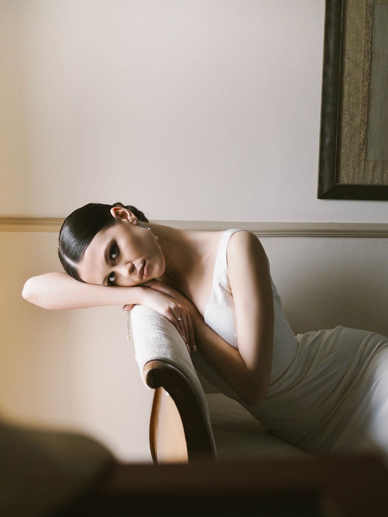 May include: A woman in a white dress is lying on a white couch with her head resting on her arm. She is looking at the camera with a thoughtful expression.