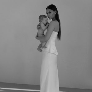 May include: A woman in a white strapless dress is holding a baby in her arms. The woman and baby are standing in front of a white wall.