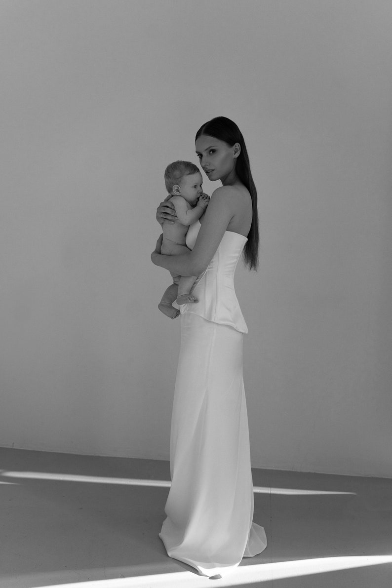 May include: A woman in a white strapless dress is holding a baby in her arms. The woman and baby are standing in front of a white wall.