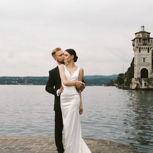 May include: A bride and groom pose for a wedding photo on a stone pier overlooking a lake. The bride is wearing a white gown with a long train and the groom is wearing a black suit. A stone tower is visible in the background.