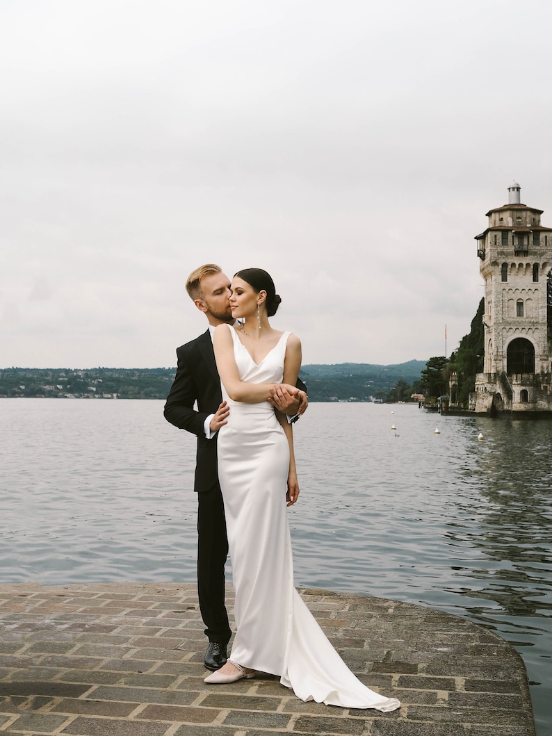 May include: A bride and groom pose for a wedding photo on a stone pier overlooking a lake. The bride is wearing a white gown with a long train and the groom is wearing a black suit. A stone tower is visible in the background.