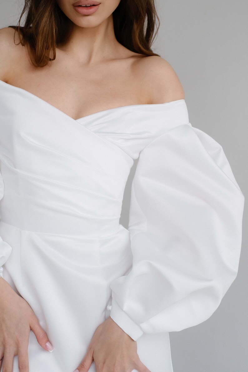 Puede incluir: Vestido blanco con hombros descubiertos y mangas largas y voluminosas. El vestido presenta un corpi&ntilde;o cruzado y una cintura entallada. Las mangas est&aacute;n recogidas en la mu&ntilde;eca. El vestido est&aacute; hecho de una tela lisa, similar al sat&eacute;n.