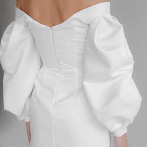 Puede incluir: Un minivestido blanco con escote de hombros descubiertos y mangas largas abullonadas. El vestido tiene un corpi&ntilde;o ajustado y una falda corta. La espalda del vestido tiene una cremallera. La tela es lisa y opaca.