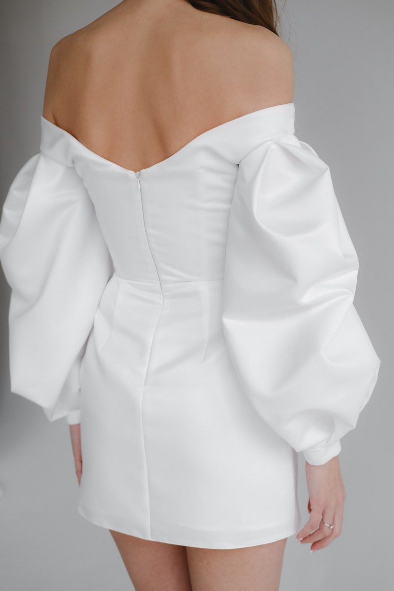 Puede incluir: Un minivestido blanco con escote de hombros descubiertos y mangas largas abullonadas. El vestido tiene un corpi&ntilde;o ajustado y una falda corta. La espalda del vestido tiene una cremallera. La tela es lisa y opaca.