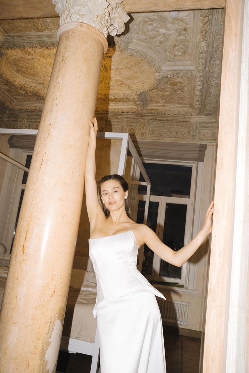 May include: A white satin corset wedding dress with a fitted bodice and a flowing skirt. The dress is shown on a mannequin against a background of an ornate interior with columns and detailed ceiling.