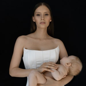 May include: A woman in a white strapless dress is holding a baby in her arms. The baby is wearing a diaper and has its eyes closed.