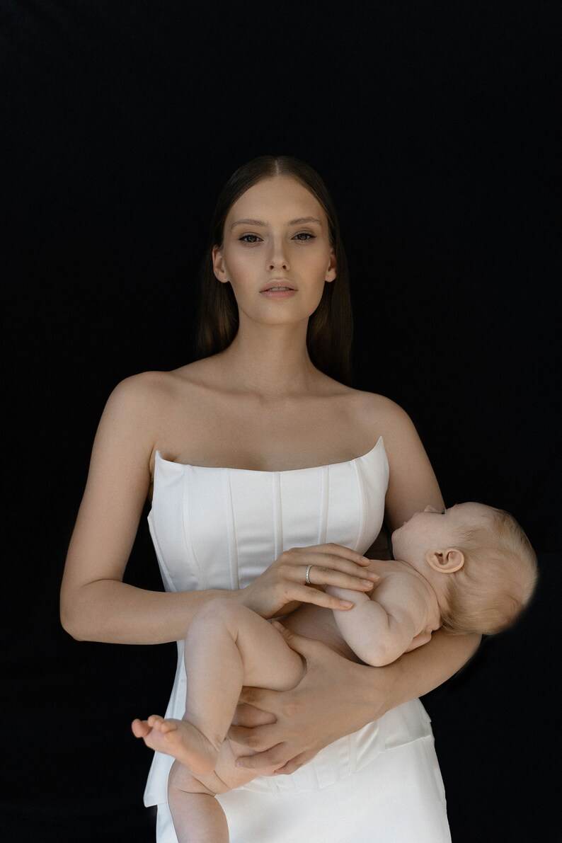 May include: A woman in a white strapless dress is holding a baby in her arms. The baby is wearing a diaper and has its eyes closed.