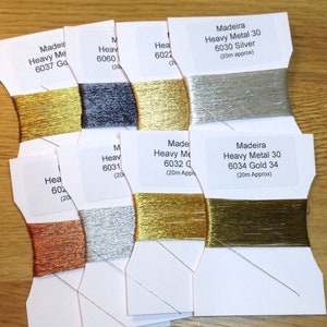 May include: Close-up of eight spools of Madeira Heavy Metal 30 embroidery thread. The spools are arranged in a row, with the thread colours visible. The colours are gold, silver, copper, and black. Each spool has a label with the Madeira brand name, the thread type, the colour number, and the length of the thread.